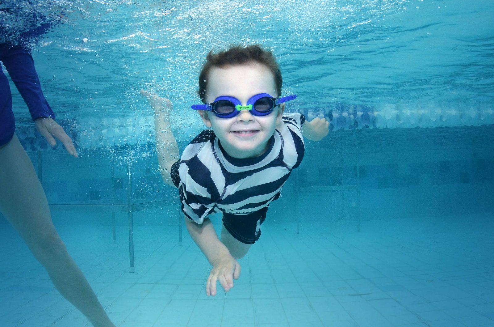 A young boy in goggles swimming underwater towards the camera during a lesson, an instructor beside him.