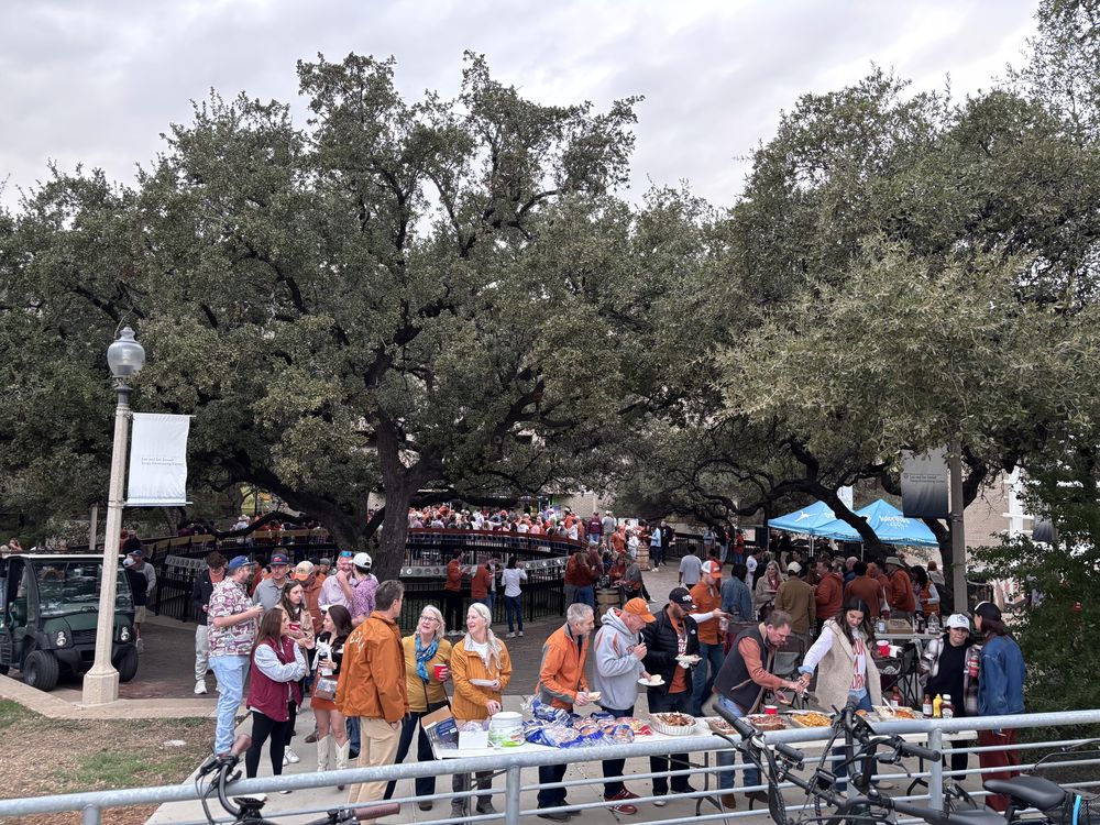 A large crowd gathered under live oak trees at the Longhorn Strokes tailgate, with food tables and sponsor tents.